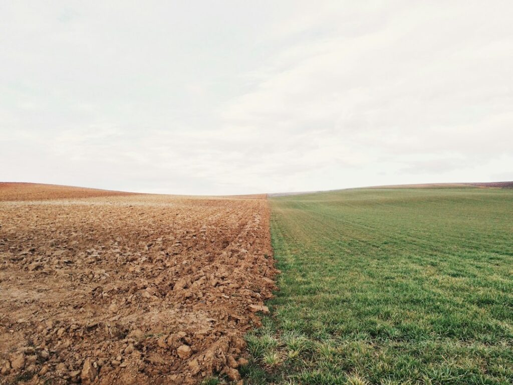 A person examines layered soil from a test pit beside a rural homesite with survey stakes and grass-covered land.